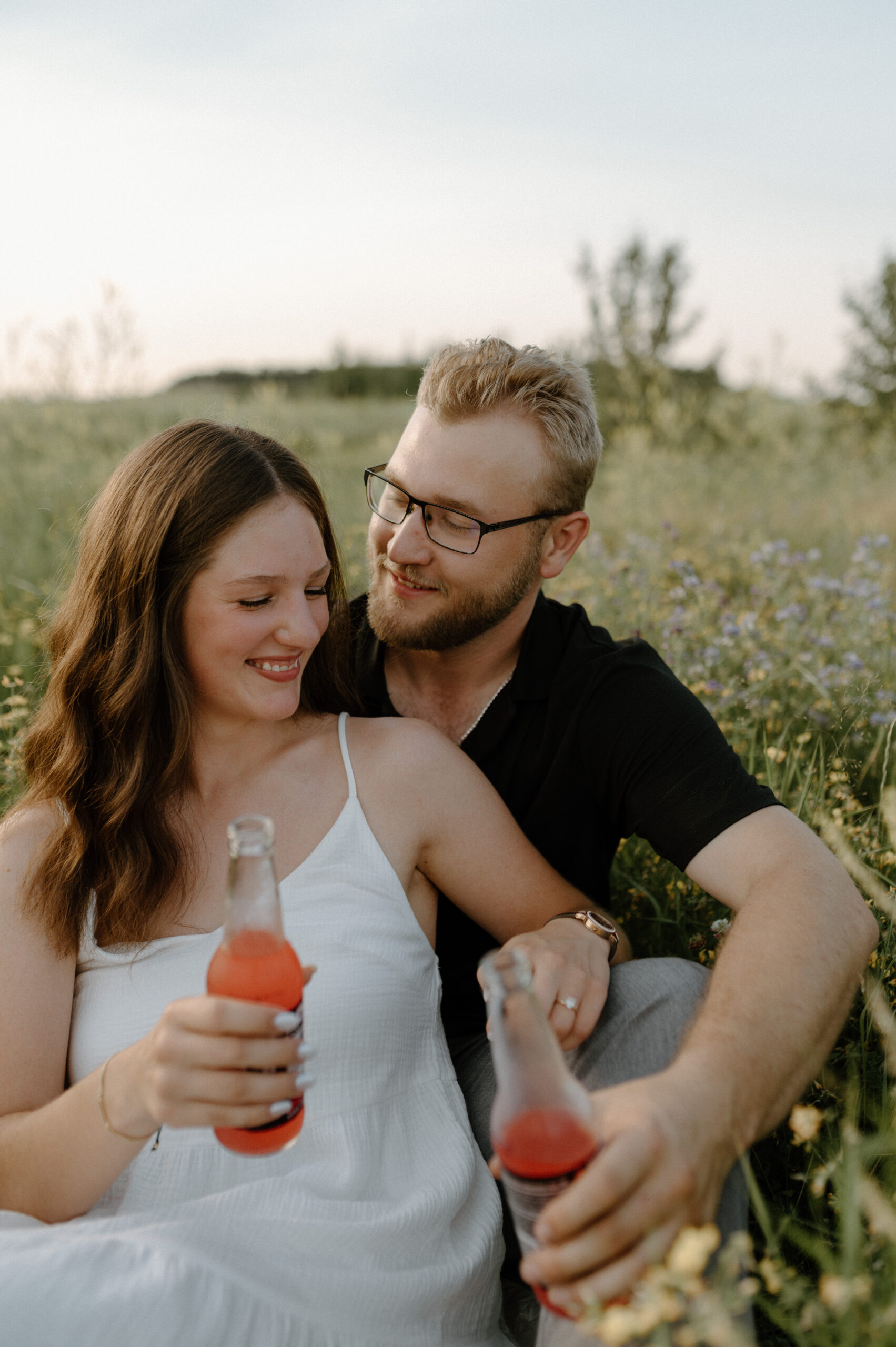 a couple sitting in tall grass and wildflowers enjoying some colourful drinks